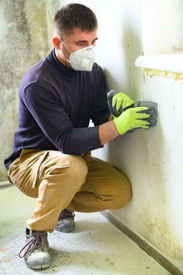 Homeowner wearing gloves and a mask while inspecting mold on a small section of drywall